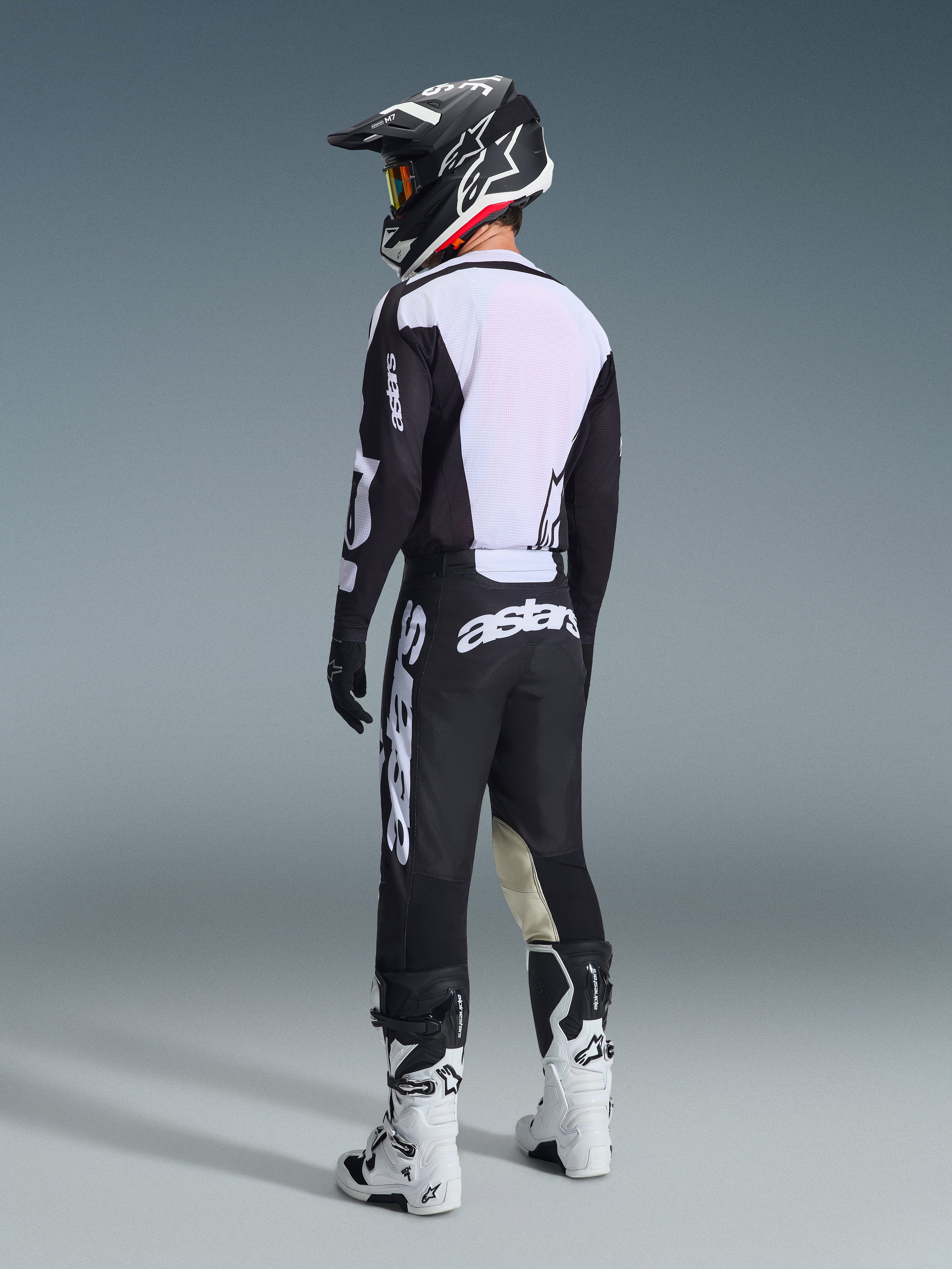 Piloto de motocross visto desde atrás con el conjunto de equipamiento Alpinestars 2026 Racer Air Riway en negro y blanco, con la espalda del jersey de malla ventilada y grandes logotipos en blanco, combinado con un casco negro a juego y botas blancas.
