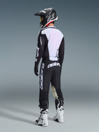 Piloto de motocross en vista posterior vistiendo el Jersey Racer Air Riway 2026 en negro y blanco, resaltando el amplio panel de malla blanca en la espalda para ventilación, con pantalones negros, casco en blanco y negro, y botas de competición blancas, equipamiento profesional completo para rendimiento off-road.