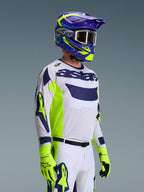 Motocross rider wearing the Alpinestars 2026 Racer Air Riway Gear Set, white jersey with navy blue logo and neon yellow side panels, white pants with matching navy and yellow accents, purple and neon yellow helmet with white goggles, neon yellow gloves, high-performance off-road racing gear