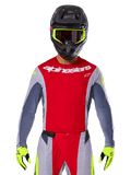 Piloto de motocross con Jersey Techstar Melt 2025 y pantalones a juego, rojo brillante y gris con logotipos de Alpinestars en blanco, casco negro con gafas amarillo neón, guantes amarillo neón a juego, equipamiento de alto rendimiento ventilado para carreras off-road