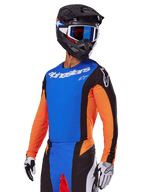 Alpinestars 2025 Techstar Melt Gear Set, piloto de motocross con jersey azul, mangas naranjas y paneles laterales negros, pantalones a juego en azul y negro, casco negro con gafas azules, guantes blancos y negros, equipación de motocross de ajuste atlético de competición