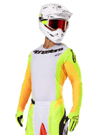 Piloto de motocross con el conjunto Alpinestars Techstar Melt 2025, jersey blanco con mangas amarillas y naranjas vibrantes, logotipo de Alpinestars negro en el pecho, guantes amarillo neón a juego, casco blanco con gafas de lente roja, kit profesional de carreras todoterreno