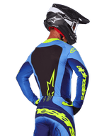 Vista posterior de un piloto de motocross con el Jersey Alpinestars Techstar Melt 2025 en azul y amarillo flúor, que presenta un panel posterior negro y llamativos logotipos amarillos en las mangas, combinado con pantalones de motocross a juego y un casco de carbono negro, equipamiento de alto rendimiento para carreras todoterreno