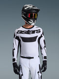 Piloto de motocross con el jersey Alpinestars Racer Riway 2026 en blanco y negro, con el logotipo 'astars' en el pecho, combinado con pantalones a juego en blanco y negro, casco negro con gafas y guantes negros, equipamiento coordinado para carreras off-road