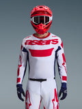 Piloto de motocross con el jersey Alpinestars 2026 Racer Riway en blanco, azul y rojo, con un gran logotipo 'astars' rojo en el pecho, paneles laterales azul marino, pantalones de motocross a juego en blanco y rojo, casco rojo con gafas blancas y guantes azul marino, conjunto completo de equipamiento para carreras todoterreno