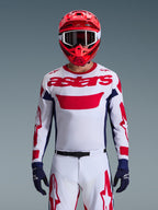 Piloto de motocross con el jersey Alpinestars 2026 Racer Riway en blanco, azul y rojo, con un gran logotipo 'astars' rojo en el pecho, paneles laterales azul marino, pantalones de motocross a juego en blanco y rojo, casco rojo con gafas blancas y guantes azul marino, conjunto completo de equipamiento para carreras todoterreno