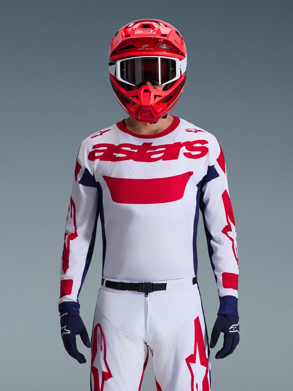 Piloto de motocross con el jersey Alpinestars 2026 Racer Riway en blanco, azul y rojo, con un gran logotipo 'astars' rojo en el pecho, paneles laterales azul marino, pantalones de motocross a juego en blanco y rojo, casco rojo con gafas blancas y guantes azul marino, conjunto completo de equipamiento para carreras todoterreno