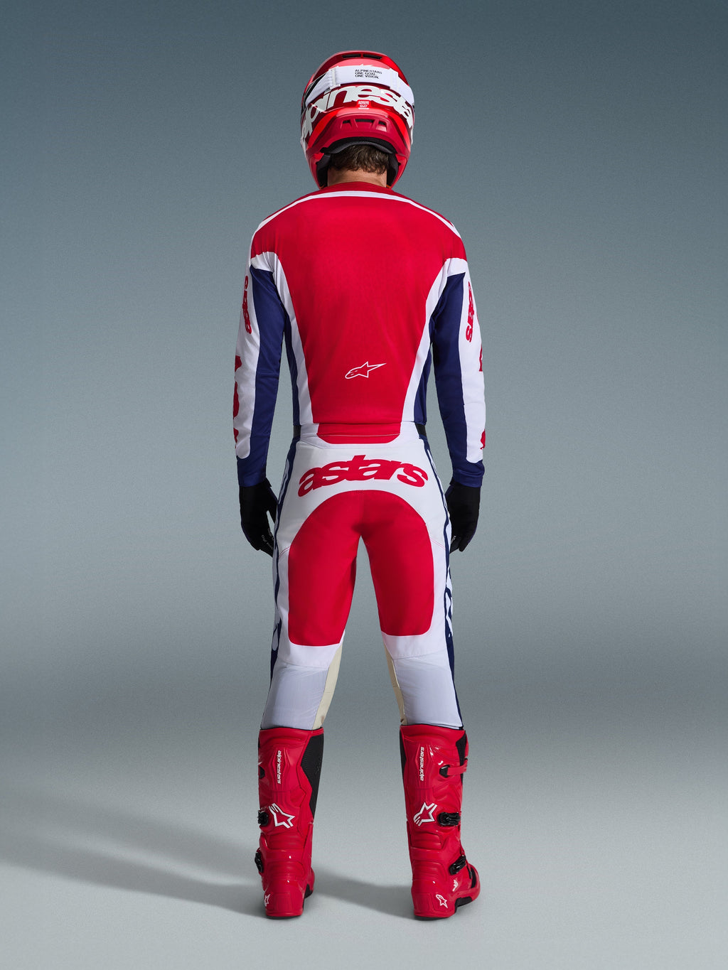Piloto de motocross visto desde atrás con el conjunto de equipación Racer Riway 2026, jersey rojo y azul con detalles blancos, pantalones blancos con paneles rojos y logo 'astars' en la parte trasera, coordinado con casco rojo, botas rojas y guantes negros, equipación de carreras de alto rendimiento para off-road