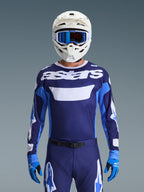 Piloto de motocross con el Jersey Alpinestars Racer Riway 2026, en azul UCLA y blanco con logotipo 'astars' blanco en el pecho, casco blanco con gafas azules, pantalones de motocross a juego y guantes azules, equipamiento de carreras off-road coordinado