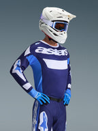 Piloto de motocross vistiendo el jersey Alpinestars 2026 Racer Riway en color UCLA Blue White, con la marca 'astars' en el pecho, combinado con pantalones de motocross azules a juego, casco blanco con gafas y guantes azules; equipamiento de alto rendimiento para carreras off-road.