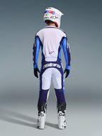 Piloto de motocross desde atrás luciendo el Jersey Alpinestars 2026 Racer Riway en azul UCLA y blanco, pantalones de motocross a juego con paneles blancos y azul marino con marca Astars, botas blancas y negras, y casco blanco, conjunto de competición coordinado para rendimiento off-road.