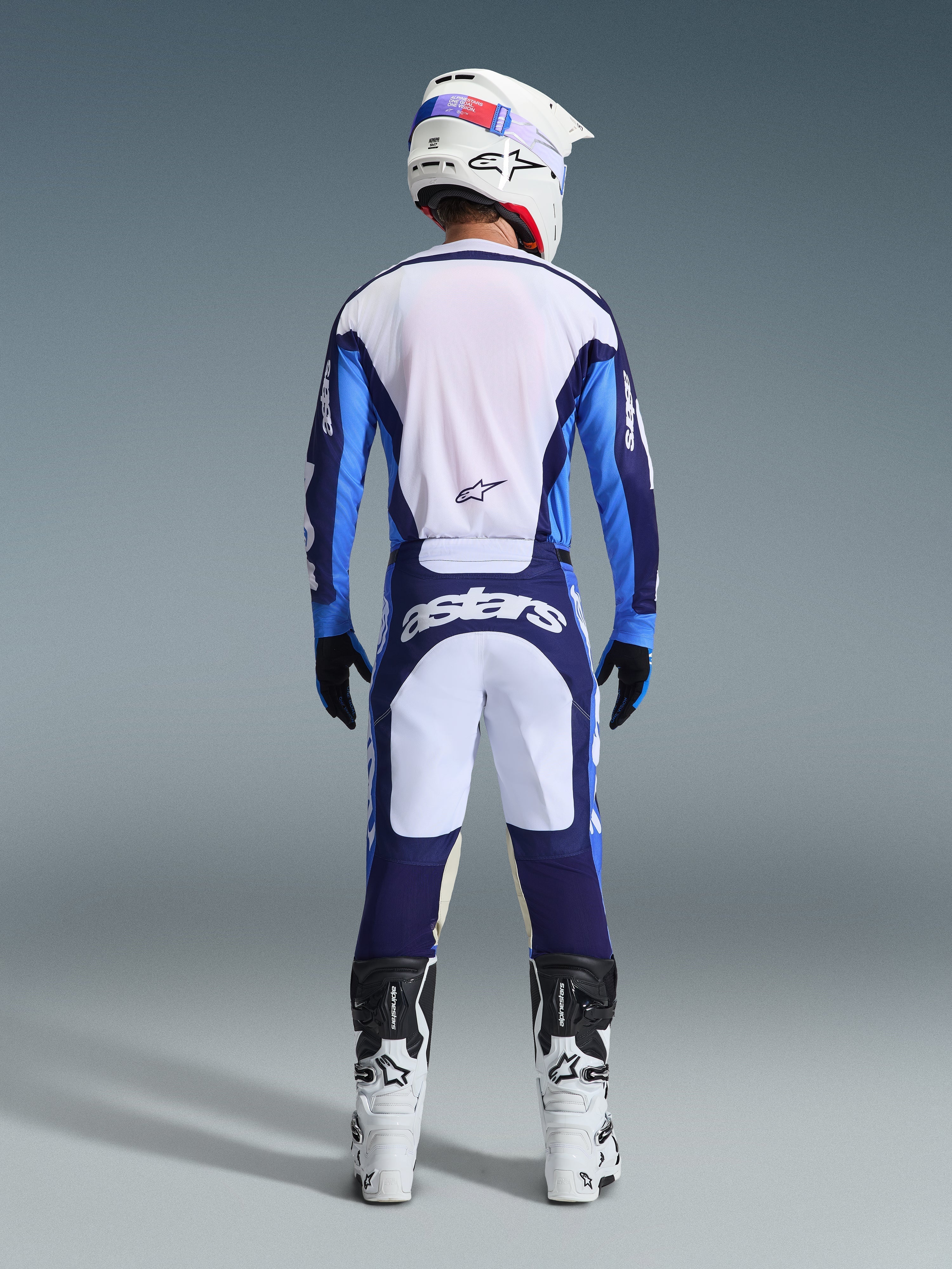 Piloto de motocross desde atrás luciendo el Jersey Alpinestars 2026 Racer Riway en azul UCLA y blanco, pantalones de motocross a juego con paneles blancos y azul marino con marca Astars, botas blancas y negras, y casco blanco, conjunto de competición coordinado para rendimiento off-road.