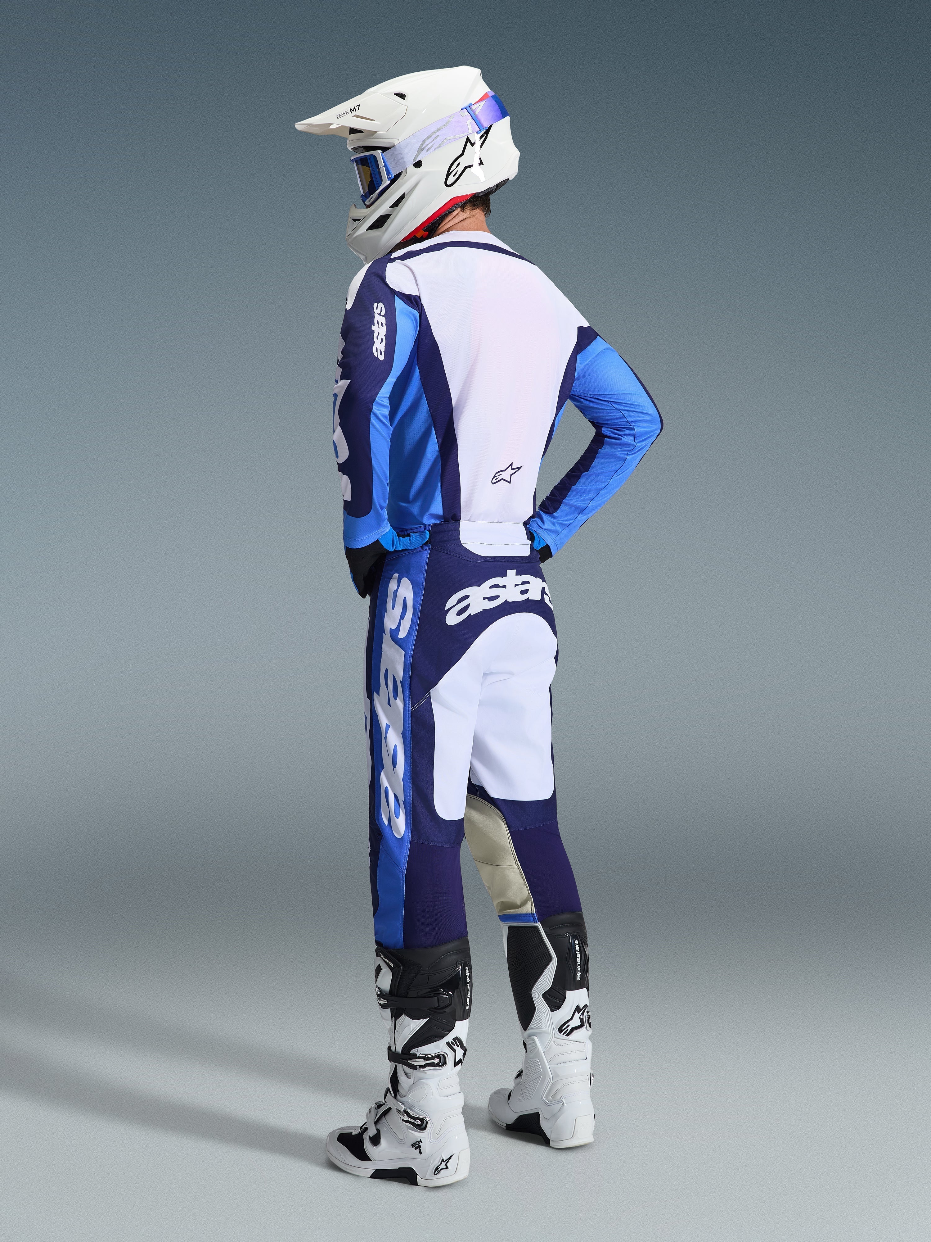 Piloto de motocross con el Jersey Racer Riway 2026 en azul Ucla y blanco, combinado con pantalones de motocross a juego en azul y blanco, casco blanco y botas blancas, en una vista de tres cuartos trasera para un look completo de carreras off-road.