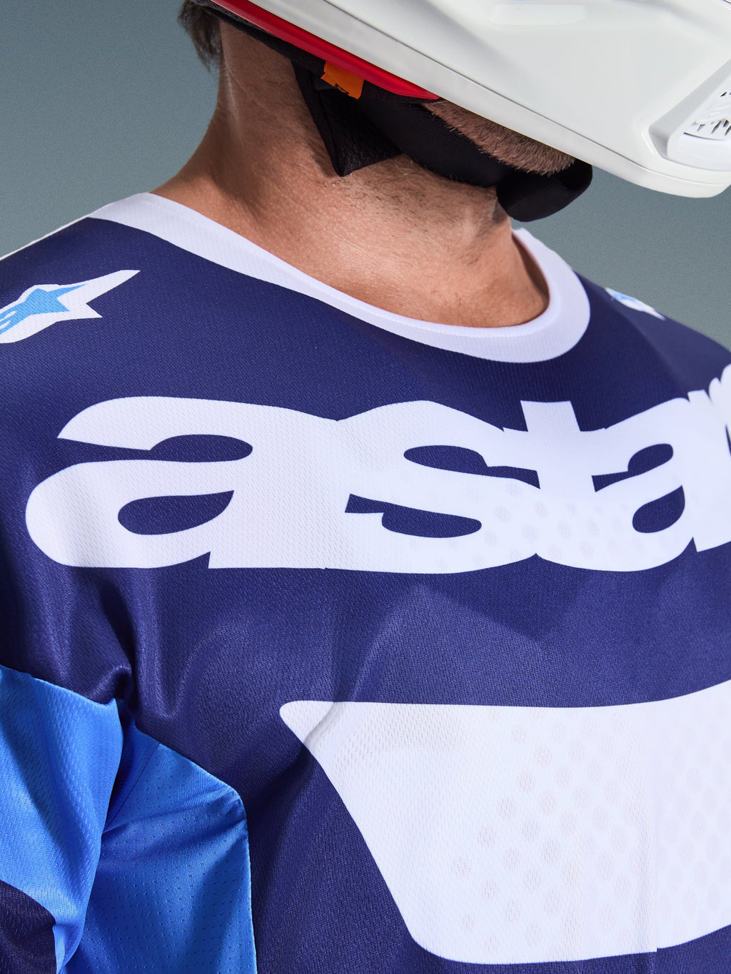 Primer plano del Jersey Alpinestars Racer Riway 2026 en azul UCLA y blanco, con un gran logotipo 'astars' blanco en el pecho, cuello blanco y textura de malla transpirable, usado con un casco de motocross blanco