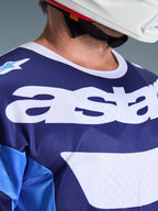 Primer plano del Jersey Alpinestars Racer Riway 2026 en azul UCLA y blanco, con un gran logotipo 'astars' blanco en el pecho, cuello blanco y textura de malla transpirable, usado con un casco de motocross blanco