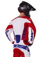 Alpinestars 2025 Racer Veil Jersey y pantalones, conjunto de equipamiento de motocross, rojo brillante, blanco y azul, vista trasera de un piloto con el equipo a juego y casco de motocross negro, con logotipos llamativos de Alpinestars en azul, ropa técnica para carreras off-road