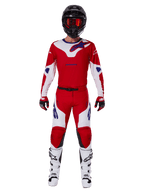 Piloto de motocross con el conjunto de equipamiento Alpinestars 2025 Racer Veil, jersey y pantalones rojo brillante y blanco con detalles azules, casco negro con gafas rojas, botas blancas y guantes rojos y negros, atuendo coordinado para carreras off-road