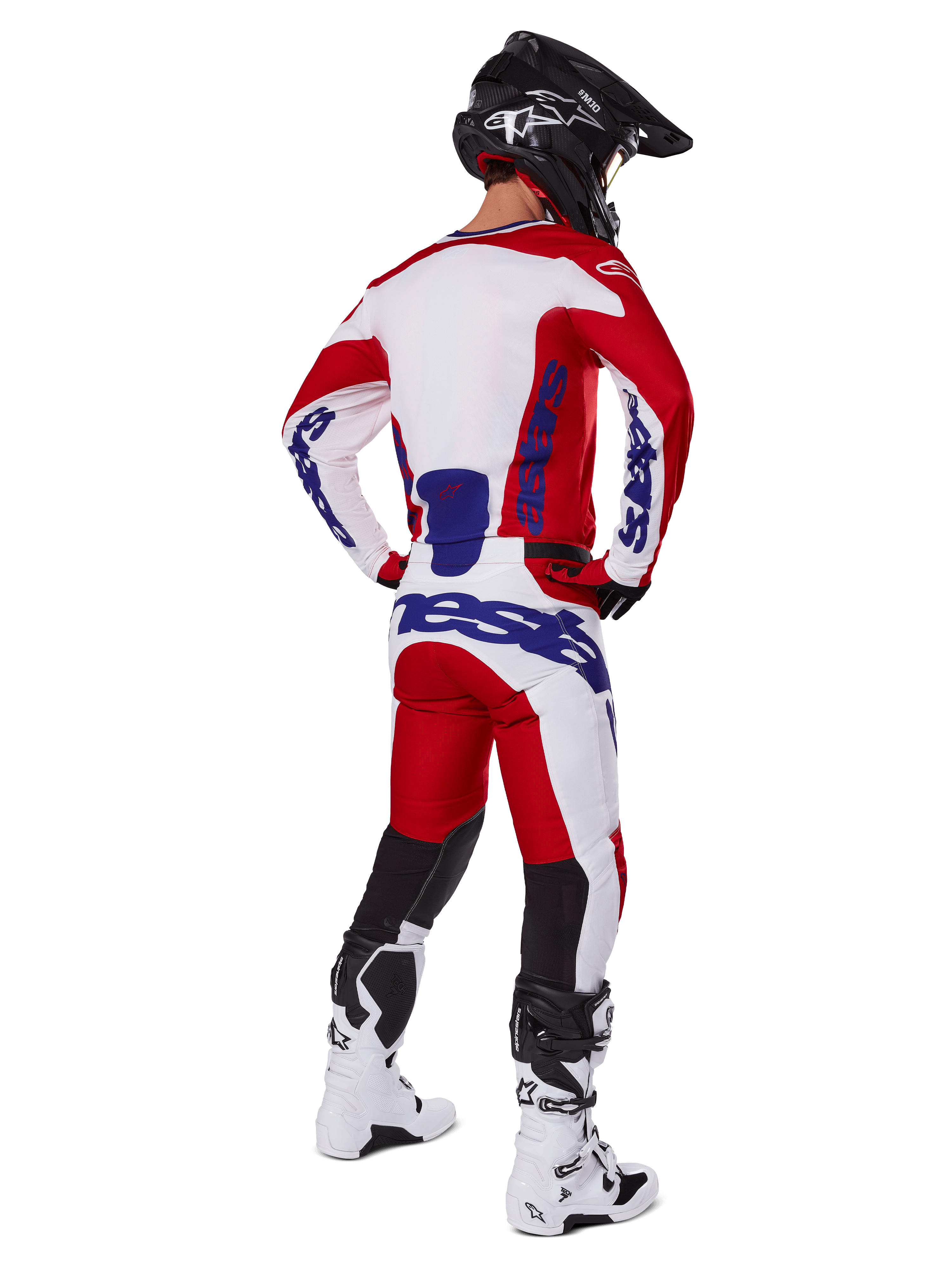 Piloto de motocross visto desde atrás vistiendo el jersey y los pantalones Alpinestars Racer Veil 2025 en rojo y blanco, con logotipos en azul, casco negro y botas blancas, conjunto completo de equipamiento para carreras off-road