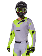 Piloto de motocross con el conjunto Alpinestars 2025 Racer Veil en gris y verde flúor, jersey y pantalones gris claro con paneles llamativos en verde neón y logotipos de Alpinestars en negro, coordinado con guantes a juego y un casco negro con gafas verde neón, equipamiento profesional para carreras todoterreno.