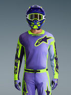 Alpinestars 2026 Racer Portl Jersey, jersey de motocross, púrpura y amarillo flúor, usado con pantalones, guantes y casco de motocross a juego, con un gran logotipo de estrella de Alpinestars en el pecho, toma de estudio sobre un fondo gris.