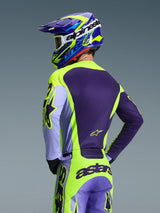 Piloto de motocross visto desde atrás con el Jersey Alpinestars 2026 Racer Portl y pantalones a juego, esquema de colores púrpura y amarillo flúor con detalles lavanda, casco coordinado en azul y amarillo, equipamiento de ajuste de carrera diseñado para el rendimiento off-road.