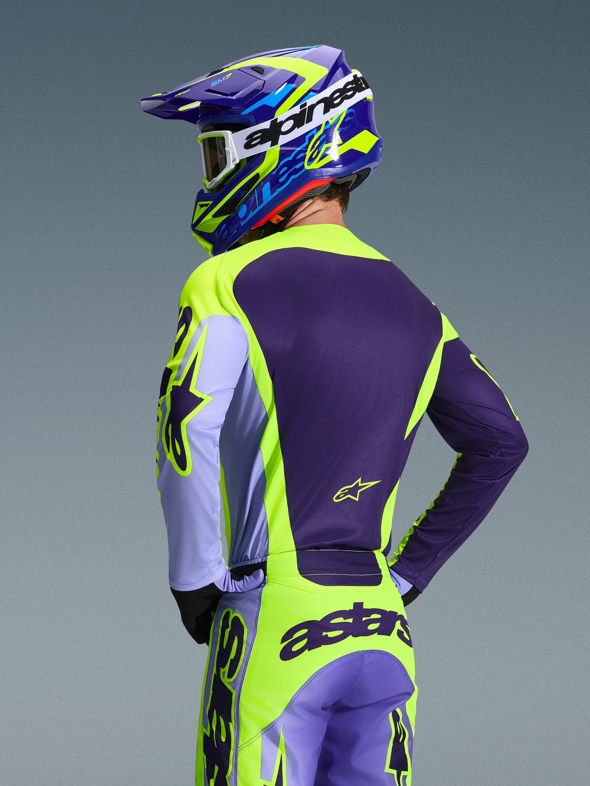 Piloto de motocross visto desde atrás con el Jersey Alpinestars 2026 Racer Portl y pantalones a juego, esquema de colores púrpura y amarillo flúor con detalles lavanda, casco coordinado en azul y amarillo, equipamiento de ajuste de carrera diseñado para el rendimiento off-road.