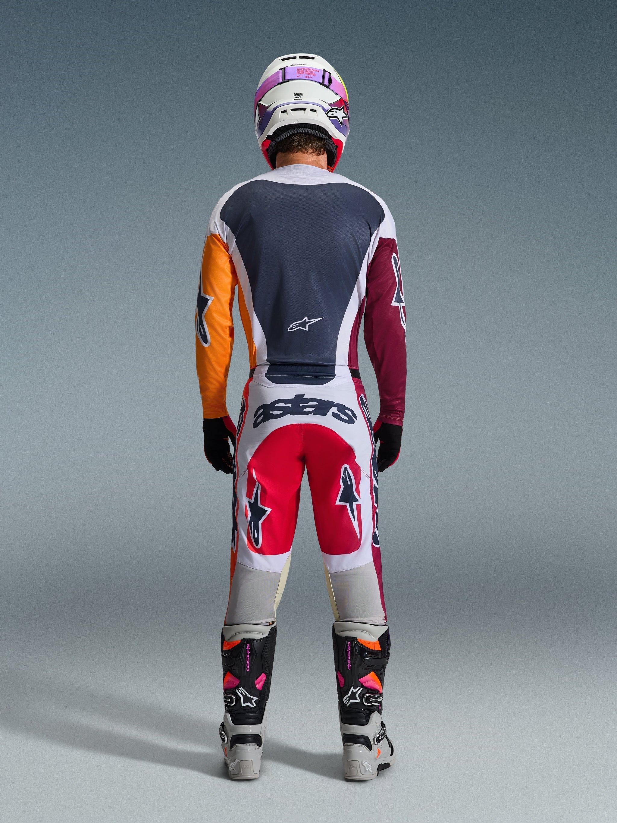 Motocross rider from behind wearing complete racing gear, 2026 Racer Portl Jersey in Red Light Gray and Dark Green with asymmetrical orange and maroon sleeves, matching white and red pants, white helmet with purple goggles, white and black boots, coordinated outfit for off-road racing