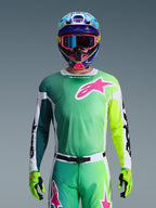 Alpinestars 2026 Racer Portl Jersey, jersey de motocross, verde, blanco y negro con un gran logotipo rosa de Alpinestars en el pecho, usado por un piloto con un casco multicolor vibrante y gafas a juego, equipamiento profesional para carreras todoterreno.