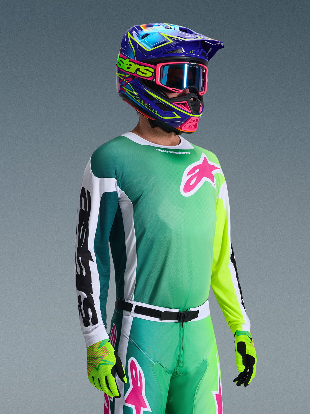 Alpinestars 2026 Racer Portl Jersey, jersey de motocross, verde, blanco y negro, usado por un piloto con pantalones verdes a juego, guantes fluorescentes y un casco azul y púrpura vibrante con gafas rosas, con un logotipo Astars rosa en el pecho.