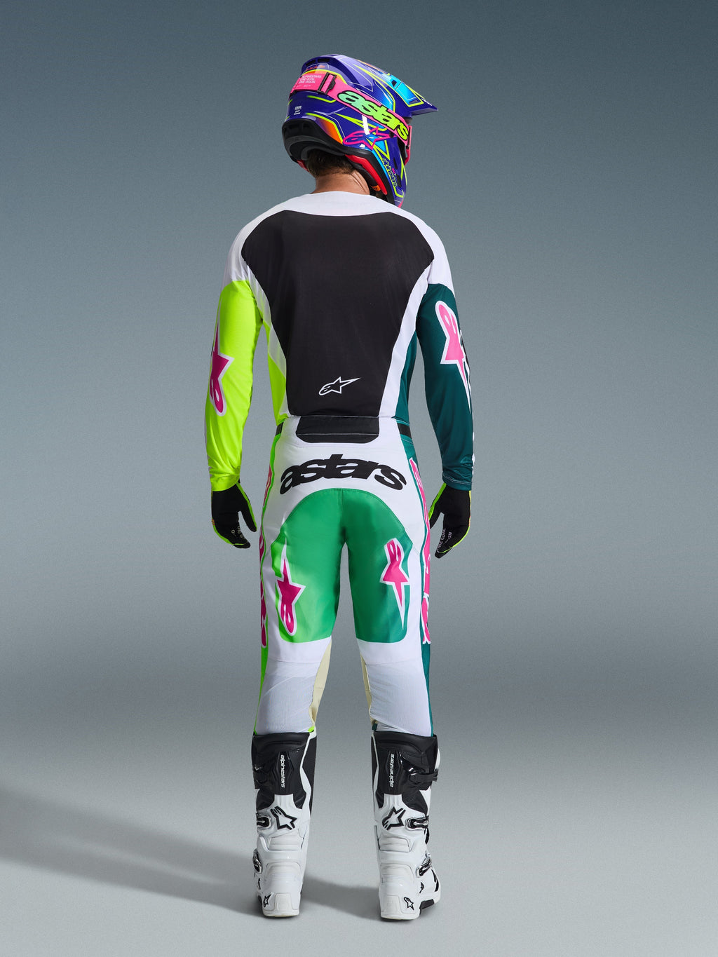 Piloto de motocross visto desde atrás luciendo el Jersey Racer Portl 2026 en color verde, blanco y negro, con un panel trasero negro y mangas verdes asimétricas con logotipos de estrellas rosas, combinado con pantalones de motocross a juego y botas blancas.