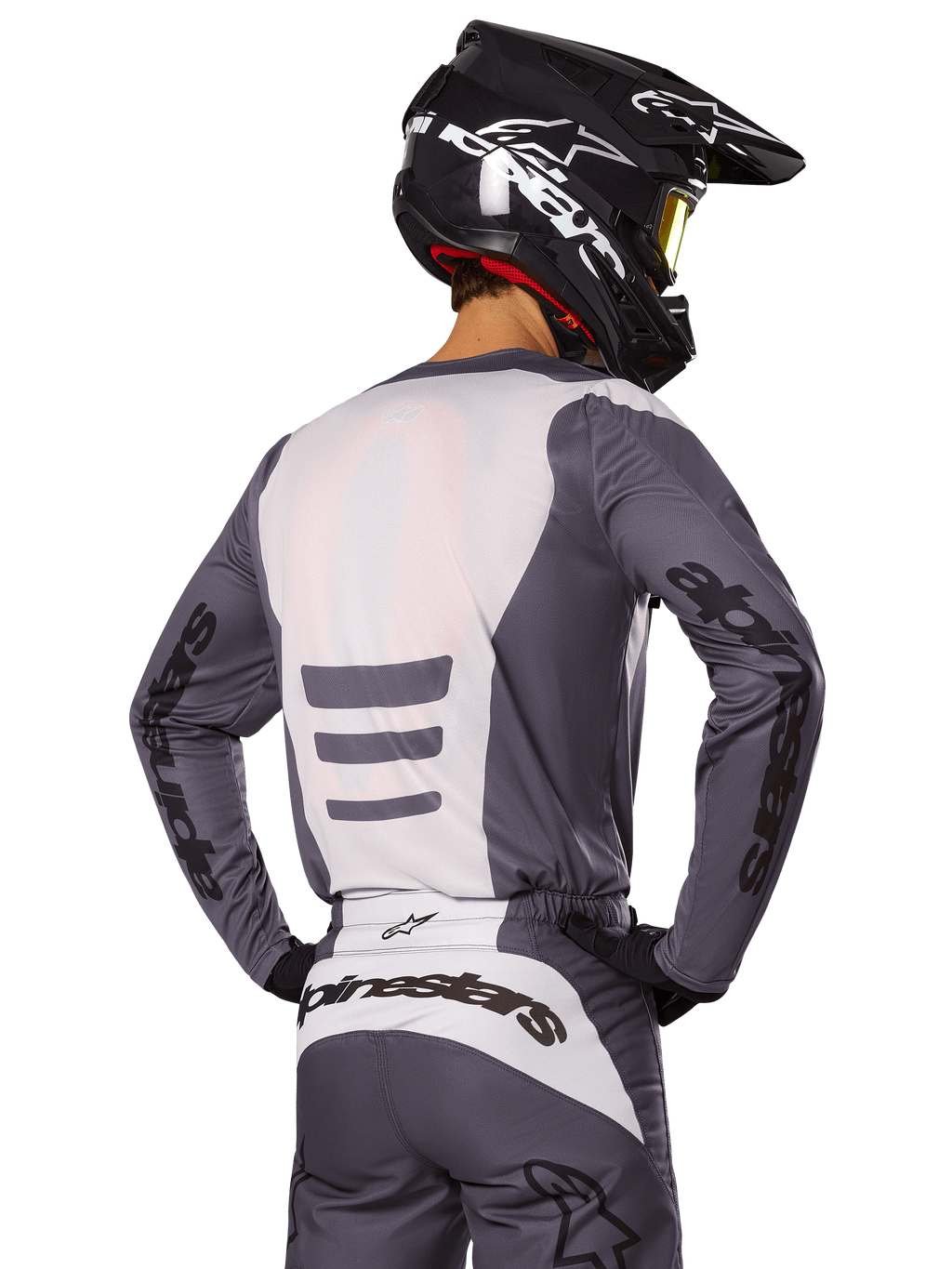 Piloto de motocross visto desde atrás con el conjunto Alpinestars 2025 Fluid Haul, jersey y pantalones en gris y blanco, casco negro con gráficos blancos, gafas amarillas, atuendo de motocross coordinado