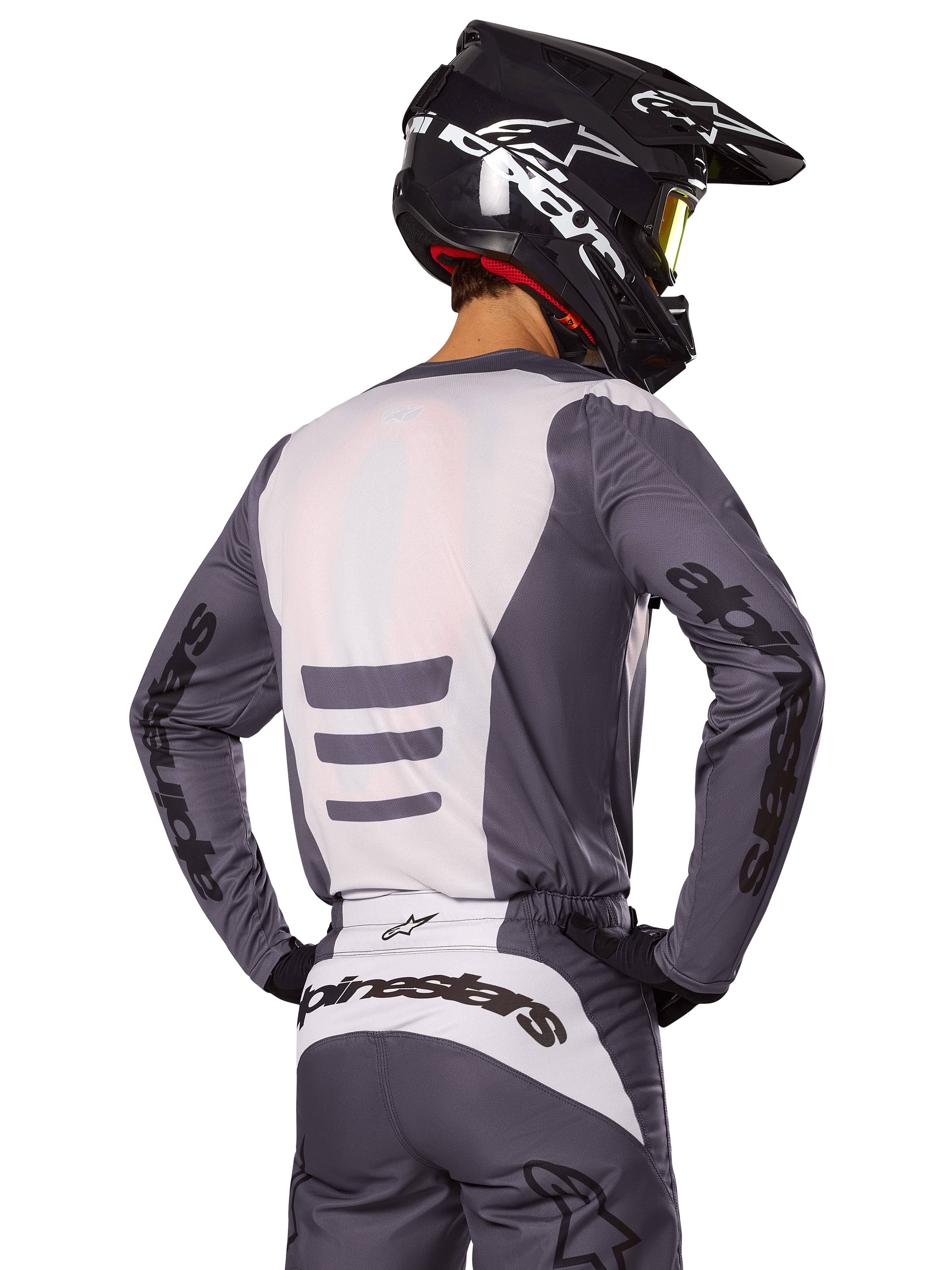 Piloto de motocross visto desde atrás con el conjunto Alpinestars 2025 Fluid Haul, jersey y pantalones en gris y blanco, casco negro con gráficos blancos, gafas amarillas, atuendo de motocross coordinado