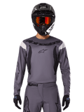 Piloto de motocross con el Jersey Fluid Haul 2025 y conjunto de equipo, gris con detalles en negro y blanco, con mangas largas y logotipos llamativos, casco negro con gafas naranjas y guantes negros, atuendo coordinado para carreras todoterreno