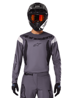 Piloto de motocross con el Jersey Fluid Haul 2025 y conjunto de equipo, gris con detalles en negro y blanco, con mangas largas y logotipos llamativos, casco negro con gafas naranjas y guantes negros, atuendo coordinado para carreras todoterreno