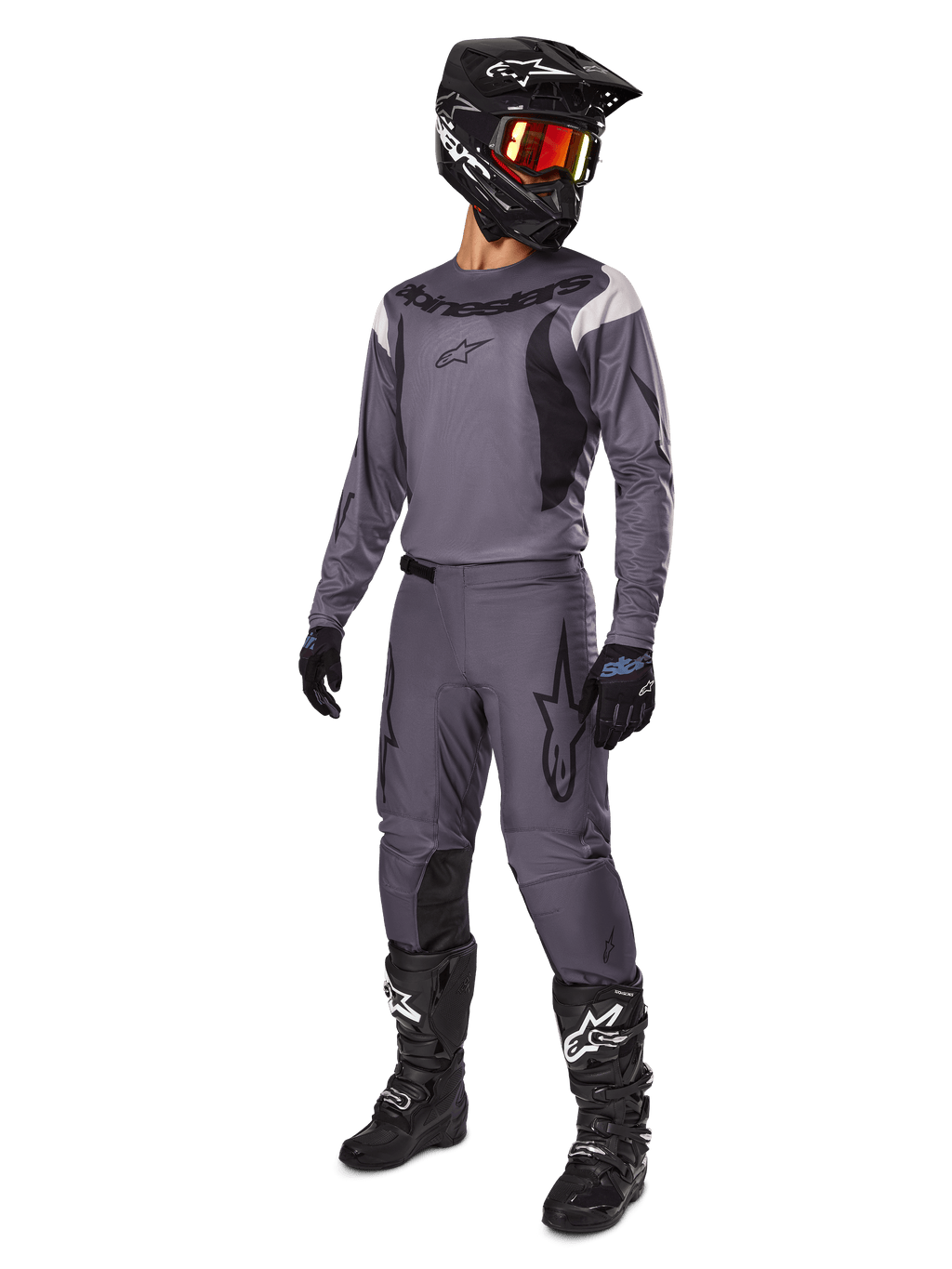 Piloto de motocross con el conjunto Alpinestars 2025 Fluid Haul en gris, que incluye jersey y pantalones grises con detalles negros, casco negro con gafas rojizas, guantes negros y botas negras con logotipos blancos, equipamiento completo para carreras todoterreno