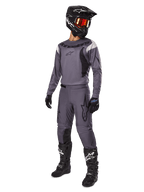 Piloto de motocross con el conjunto Alpinestars 2025 Fluid Haul en gris, que incluye jersey y pantalones grises con detalles negros, casco negro con gafas rojizas, guantes negros y botas negras con logotipos blancos, equipamiento completo para carreras todoterreno