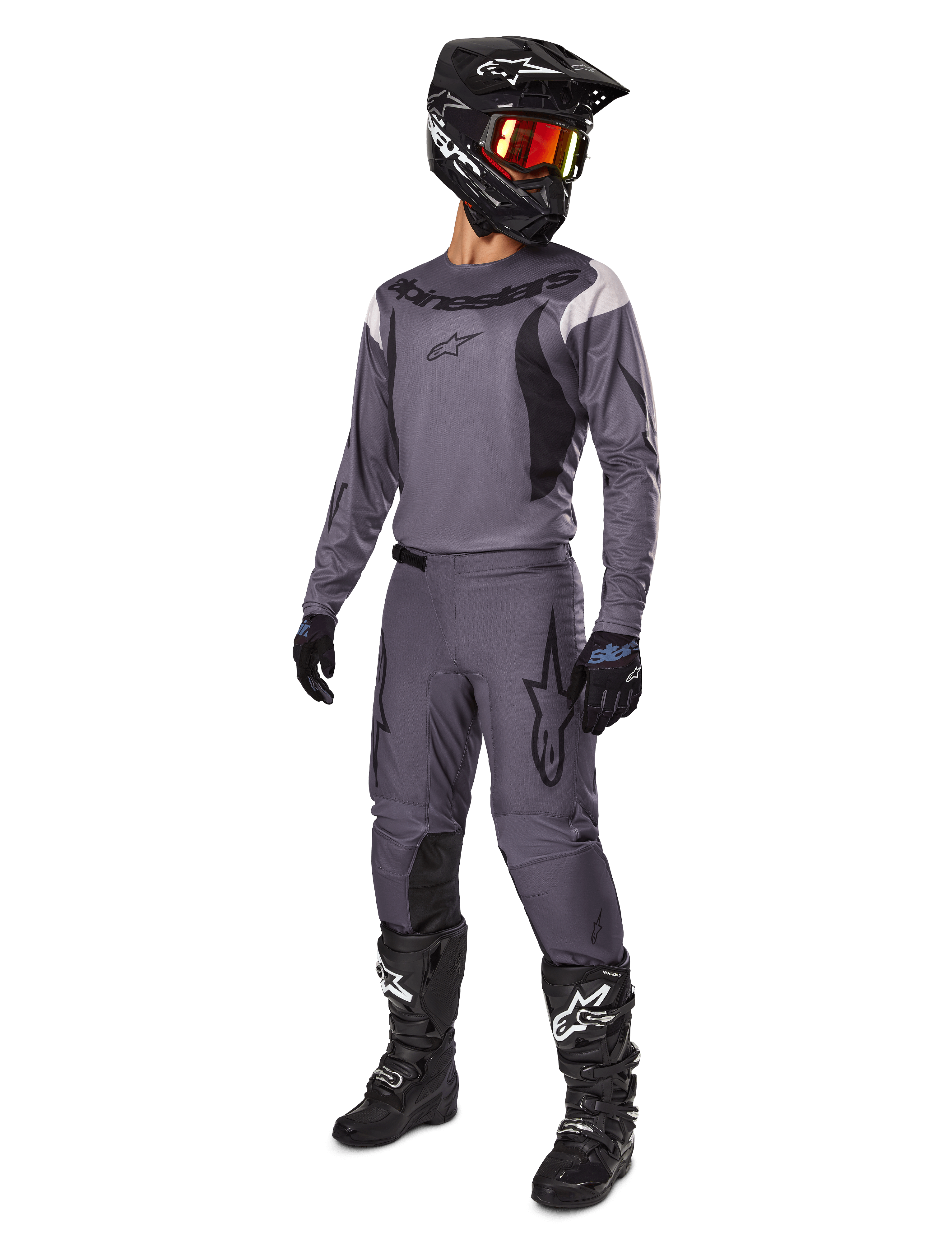 Piloto de motocross con el conjunto Alpinestars 2025 Fluid Haul en gris, que incluye jersey y pantalones grises con detalles negros, casco negro con gafas rojizas, guantes negros y botas negras con logotipos blancos, equipamiento completo para carreras todoterreno