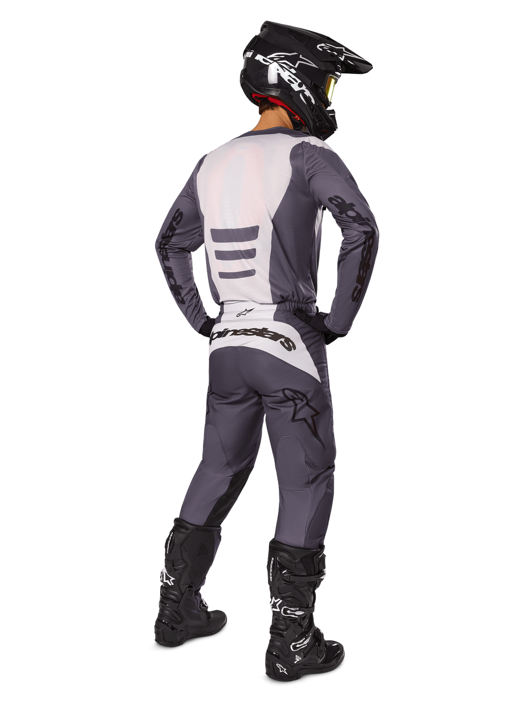 Piloto de motocross visto desde atrás vistiendo el conjunto de equipamiento Alpinestars 2025 Fluid Haul, jersey gris y blanco con pantalones a juego, casco negro con gafas amarillas, botas de motocross negras, equipamiento de competición completo para rendimiento off-road