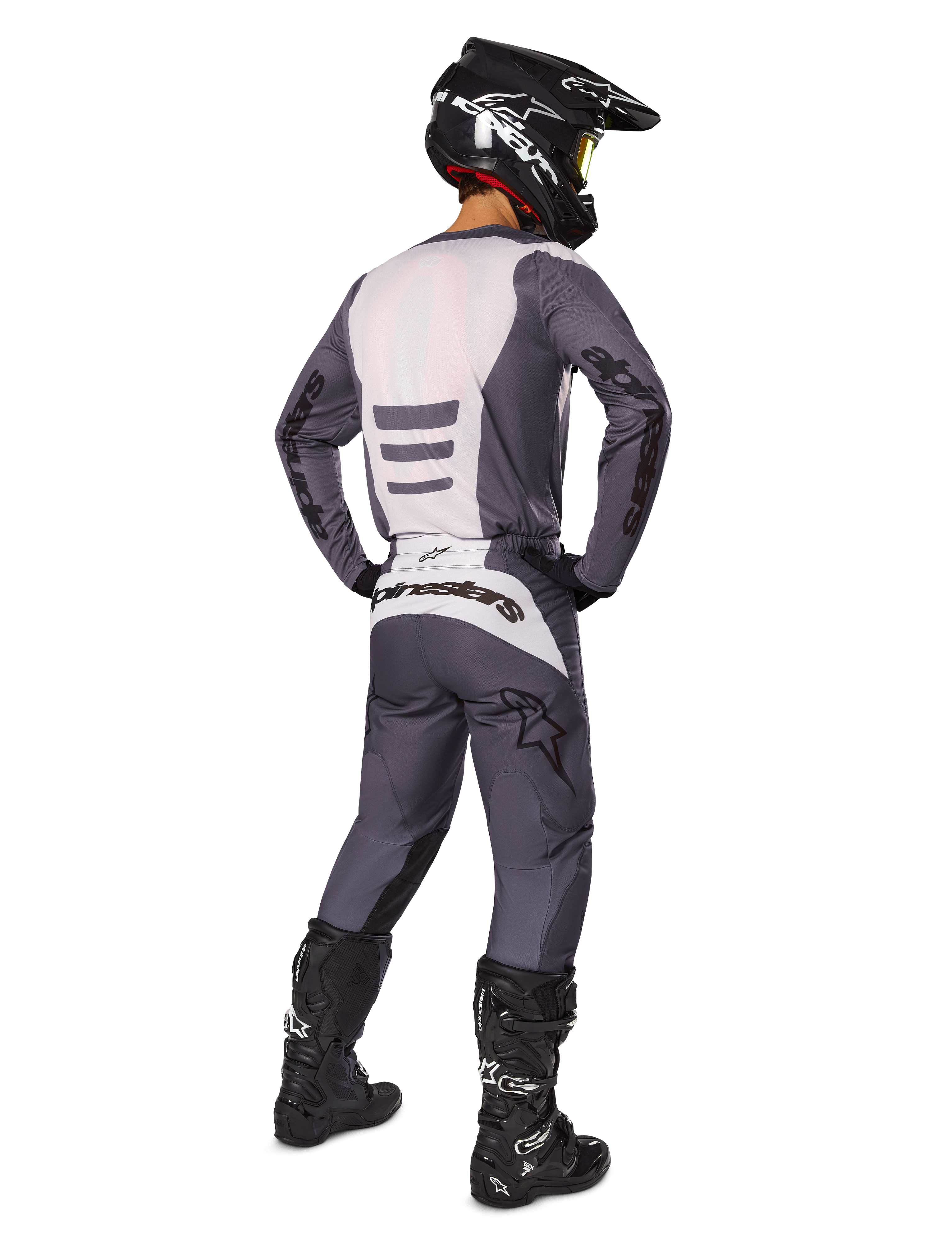 Piloto de motocross visto desde atrás vistiendo el conjunto de equipamiento Alpinestars 2025 Fluid Haul, jersey gris y blanco con pantalones a juego, casco negro con gafas amarillas, botas de motocross negras, equipamiento de competición completo para rendimiento off-road