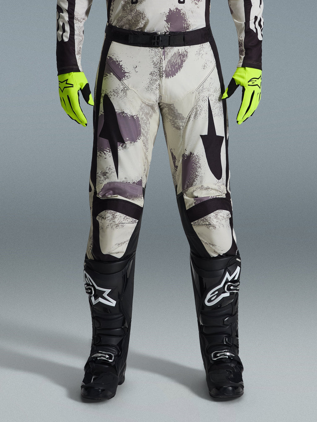 Alpinestars 2026 Racer Lahnd Pants, pantalones de motocross, patrón Sand Camo con gráficos abstractos en gris y morado, usados con guantes amarillo neón y botas negras, con logotipos de estrellas negras en los muslos y un cinturón negro, equipamiento duradero para carreras off-road