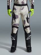 Alpinestars 2026 Racer Lahnd Pants, pantalones de motocross, patrón Sand Camo con gráficos abstractos en gris y morado, usados con guantes amarillo neón y botas negras, con logotipos de estrellas negras en los muslos y un cinturón negro, equipamiento duradero para carreras off-road