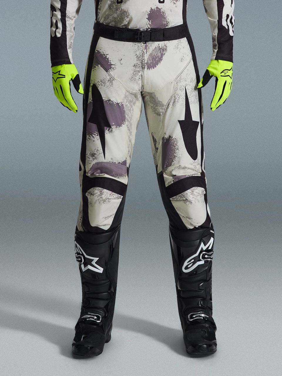 Alpinestars 2026 Racer Lahnd Pants, Motocross Pants, Sand Camo pattern with gray and purple abstract graphics, worn with neon yellow gloves and black boots, featuring black star logos on the thighs and a black waist belt, durable off-road racing gear