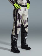 Alpinestars 2026 Racer Lahnd Pants, pantalones de motocross, Sand Camo, usados con guantes amarillo neón y botas de motocross negras, con un patrón de camuflaje abstracto y paneles laterales negros, diseño ergonómico off-road