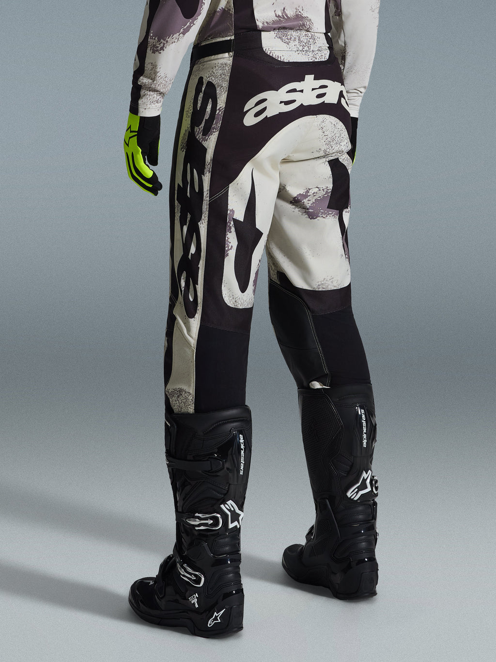 Alpinestars 2026 Racer Lahnd Pants, pantalones de motocross, camuflaje arena y gris, vista trasera con marca blanca destacada en el asiento y las piernas, combinados con botas de motocross negras y guantes amarillos, equipamiento profesional para carreras todoterreno