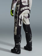 Alpinestars 2026 Racer Lahnd Pants, pantalones de motocross, camuflaje arena y gris, vista trasera con marca blanca destacada en el asiento y las piernas, combinados con botas de motocross negras y guantes amarillos, equipamiento profesional para carreras todoterreno