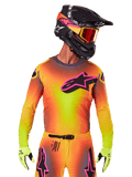 Piloto de motocross con el Jersey Supertech Lipan 2025, degradado vibrante en amarillo, naranja y rosa, gran logotipo de estrella de Alpinestars negro con contorno rosa en el pecho, perforaciones cortadas con láser para ventilación, ajuste atlético de competición, combinado con pantalones a juego, guantes amarillos y casco negro con gafas.