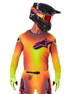 Piloto de motocross con el Jersey Supertech Lipan 2025, degradado vibrante en amarillo, naranja y rosa, gran logotipo de estrella de Alpinestars negro con contorno rosa en el pecho, perforaciones cortadas con láser para ventilación, ajuste atlético de competición, combinado con pantalones a juego, guantes amarillos y casco negro con gafas.