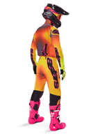 Piloto de motocross desde atrás con el conjunto de equipo Alpinestars 2025 Supertech Lipan, jersey con degradado amarillo y naranja con detalles rosas, pantalones a juego, botas rosa brillante y casco negro con gráficos en rosa y amarillo, atuendo coordinado para carreras profesionales