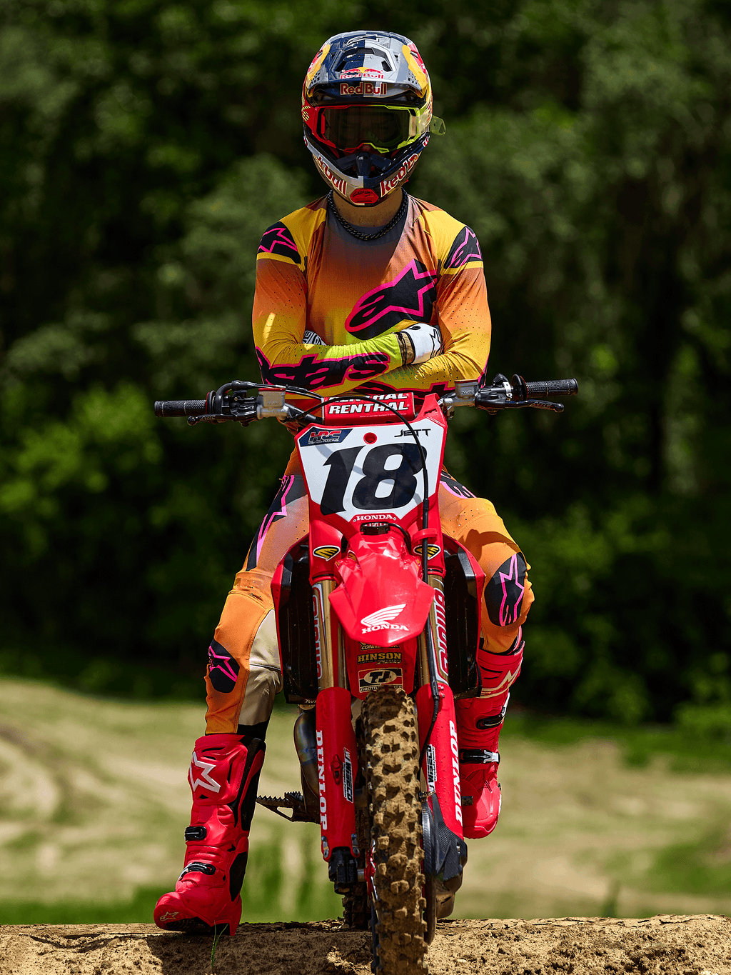 Piloto de motocross sentado en una moto Honda roja con el número 18, vistiendo el jersey Alpinestars Supertech Lipan 2025 y pantalones a juego en amarillo y rosa, botas Alpinestars rosa brillante, casco Red Bull, equipo de competición profesional para rendimiento todoterreno.
