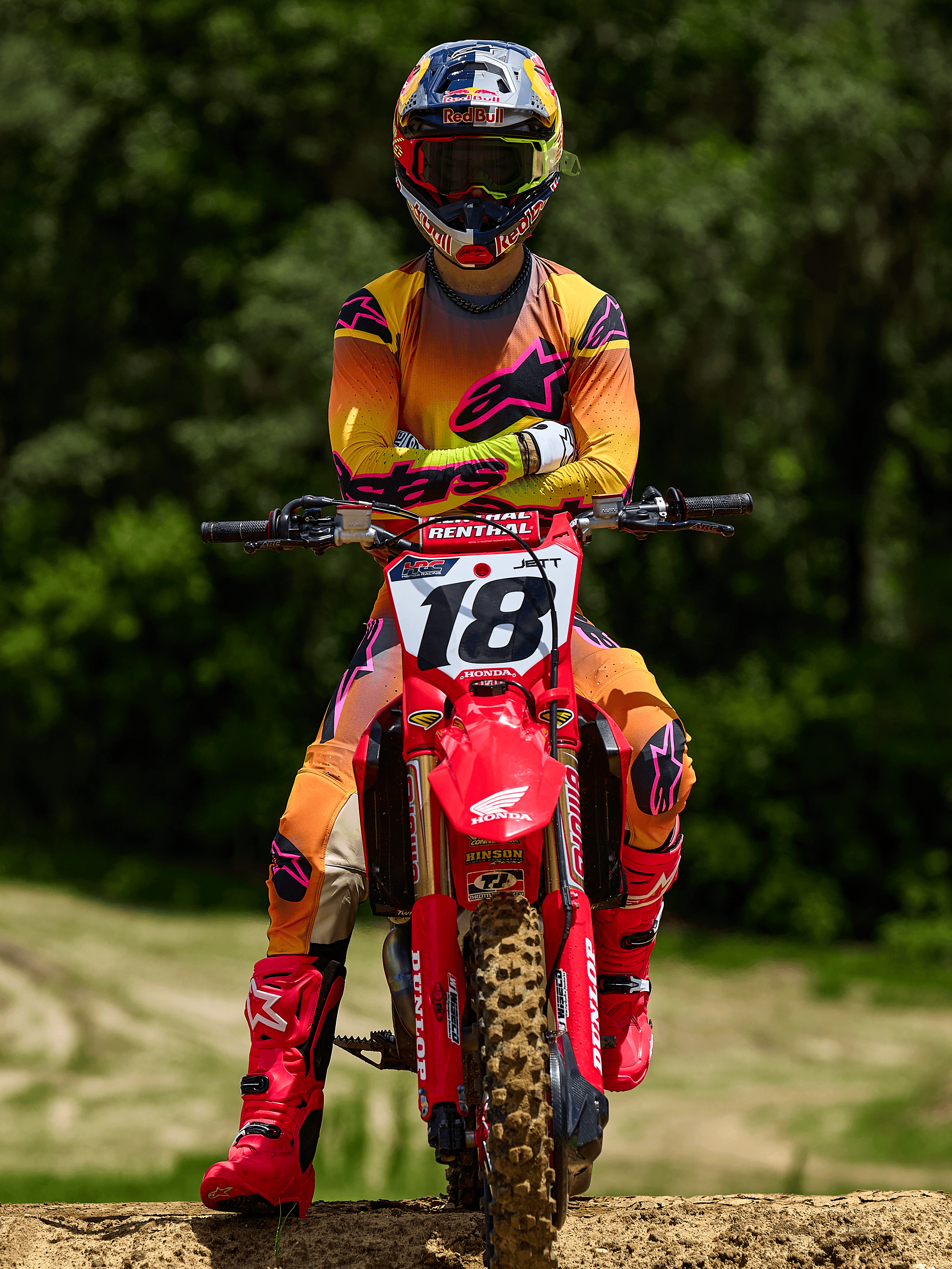 Piloto de motocross sentado en una moto Honda roja con el número 18, vistiendo el jersey Alpinestars Supertech Lipan 2025 y pantalones a juego en amarillo y rosa, botas Alpinestars rosa brillante, casco Red Bull, equipo de competición profesional para rendimiento todoterreno.